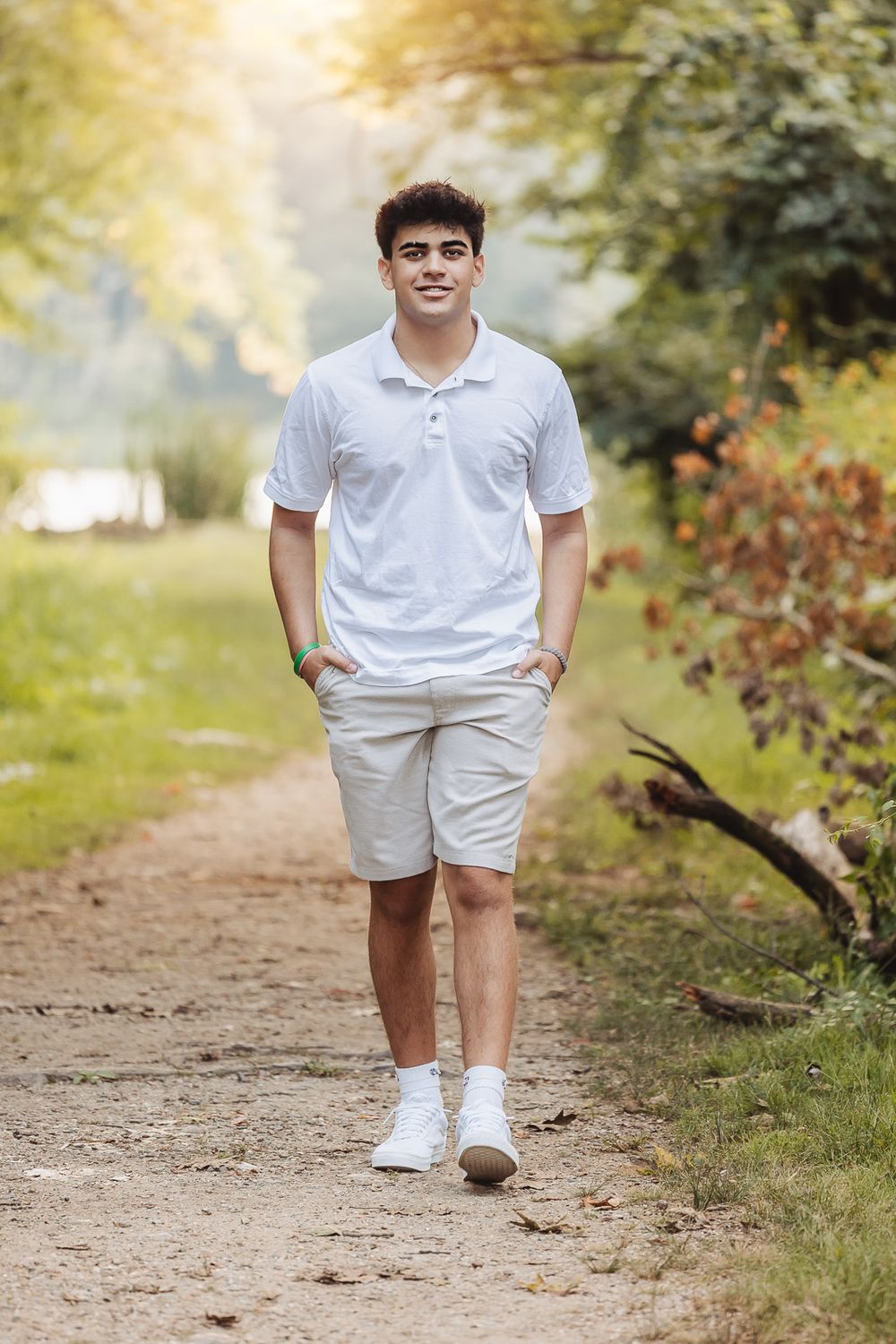 Young person in casual white polo and khaki shorts standing on a dirt path in a sunlit outdoor setting with trees and foliage.