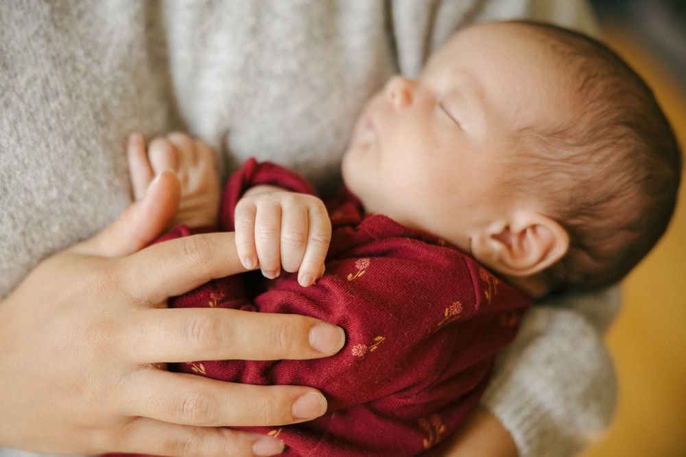 Sleeping baby in red clothing rests peacefully while being cradled by an adult hand on soft blanket.