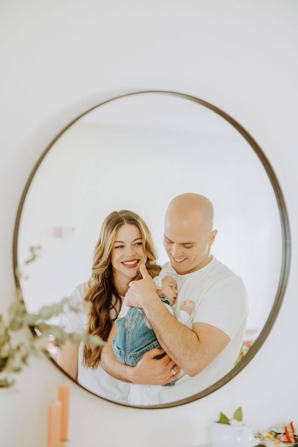 Three oval mirrors on white wall reflect intimate moments between a couple spending time with their baby.