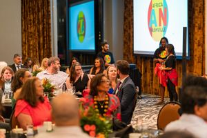 Event attendees seated at round tables during a presentation with a projection screen visible in the background.