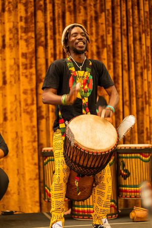 Performer in colorful African attire plays traditional djembe drum with enthusiasm on stage.