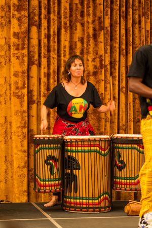 Performer in red skirt playing traditional African drums against gold curtain backdrop during cultural performance.