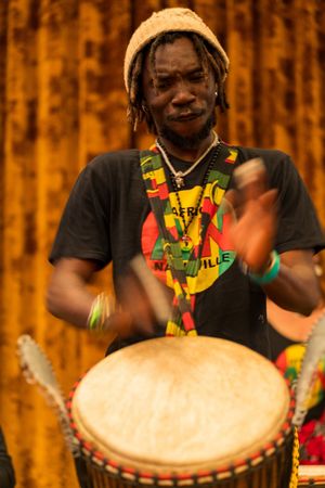 A drummer in traditional African attire plays djembe drum against an orange curtain backdrop.