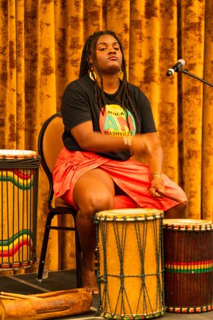 A musician in black plays traditional drums against a warm orange curtain backdrop during a performance.