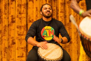Musician plays traditional African drum with joyful expression against orange curtain.