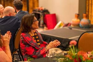 Woman in a vibrant red jacket sits attentively at a formal event with decorative centerpieces.