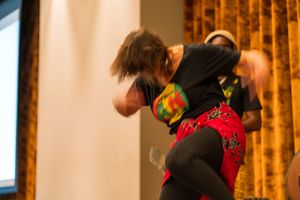 Performers in colorful attire dancing energetically on stage with curtains in the background.