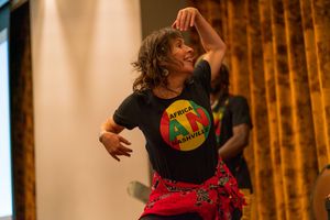 Performer in black t-shirt and red skirt dances energetically against orange curtain backdrop.