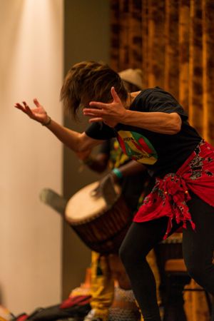 Performer in black and red outfit dances expressively with drums in dimly lit stage setting.