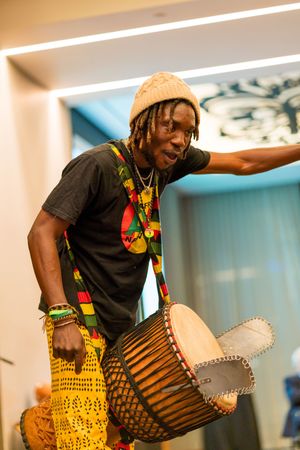 Musician in beanie and colorful necklace plays traditional African drum during performance.