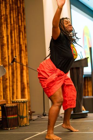 Dancer performs expressive movement in black shirt and red shorts on stage with gold curtains in background.