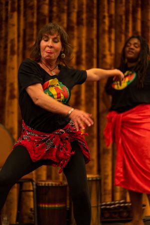 Dancers in black shirts and red garments perform energetic moves in front of a wooden bamboo wall.