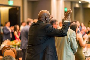 Man in suit raises his hand while standing among audience members at an evening celebration.