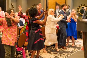 A diverse group of enthusiastic people worship together at a church gathering, raising their hands in praise and celebration.