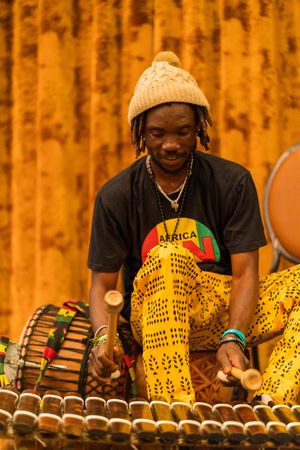 Musician in winter hat playing traditional African xylophone instrument against orange backdrop.