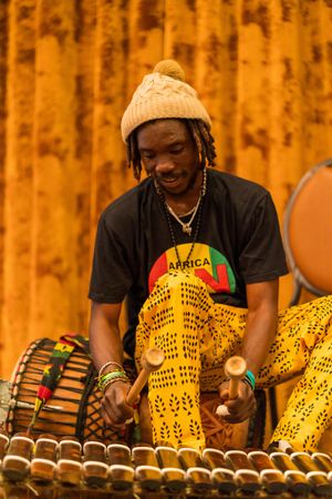 Musician in knit hat playing wooden percussion instrument against golden curtain backdrop.
