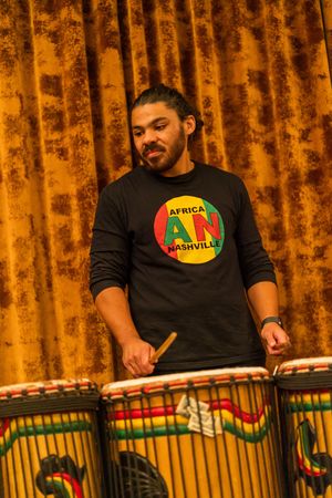 Musicians performing with traditional African drums on stage with orange lighting and decorative backdrop.
