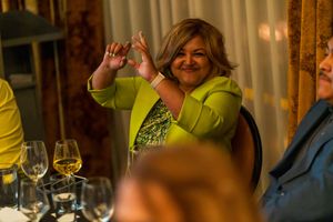 Someone in a bright yellow shirt making a heart gesture while seated at a formal dining table.