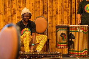 Performer in patterned yellow pants playing xylophone with African drums in the background.