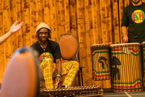 African drummer in colorful attire performs with traditional drums against orange curtain backdrop.