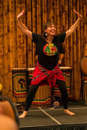 An energetic dancer performs in a traditional African-inspired outfit with colorful drums in the background.