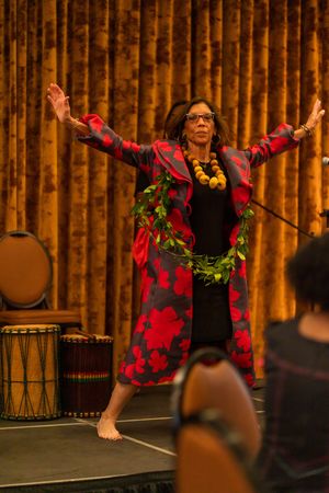 A performer in a vibrant red and black robe with a holiday wreath gestures expressively on stage with drums nearby.