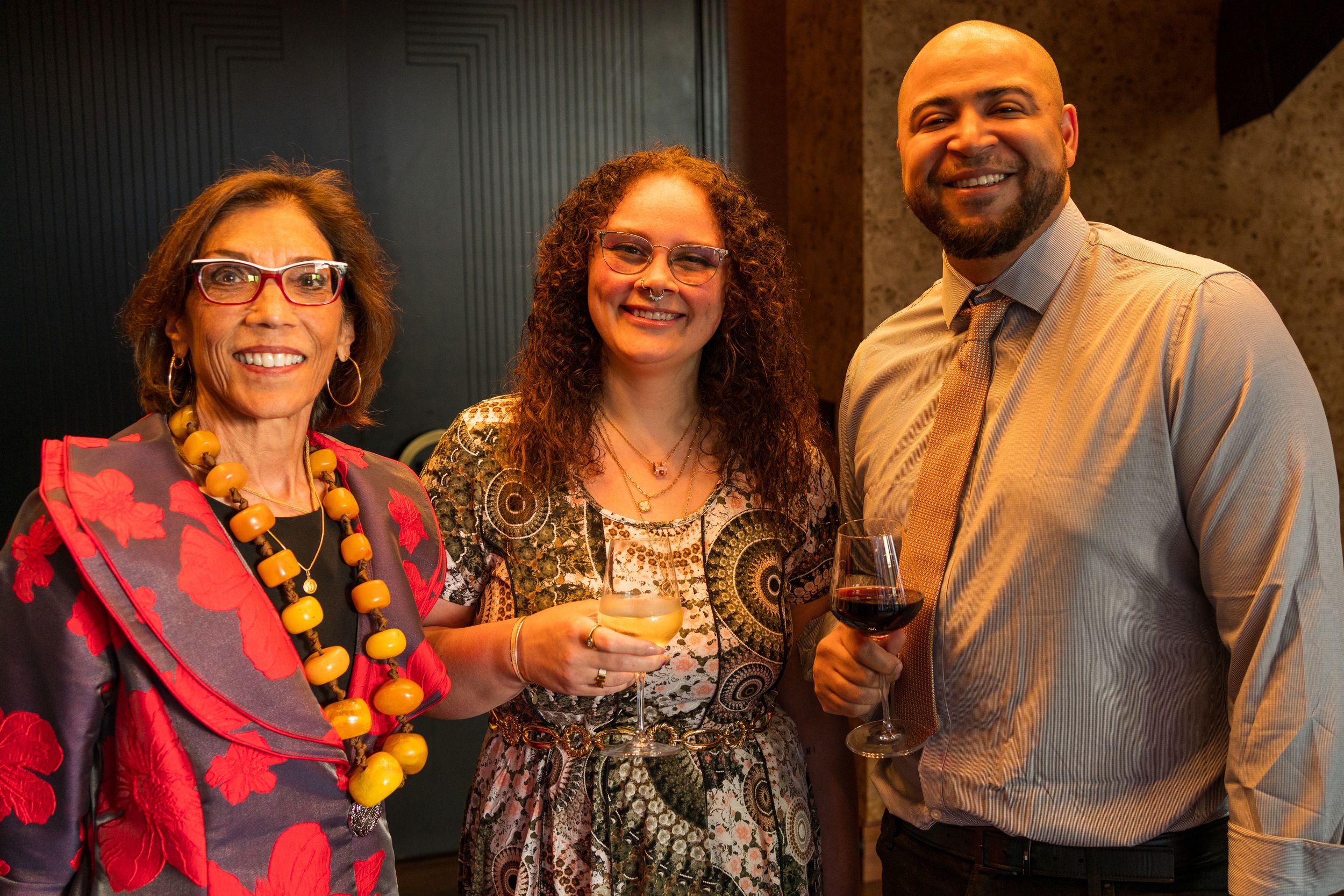 Three people in festive attire smile together at an indoor social gathering.