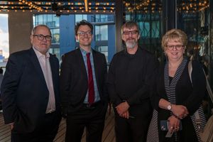 Four professionally dressed people standing together at an evening event with string lights in the background.