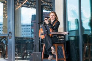 Musician performing on acoustic guitar while seated on stool at outdoor venue with city views.