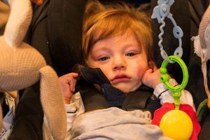 Young child in stroller looking tired with colorful toys hanging nearby.