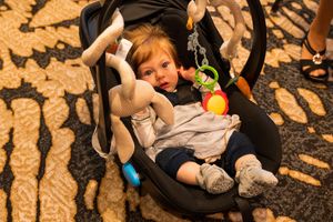 Young child in car seat reaching for colorful toy during outdoor event.