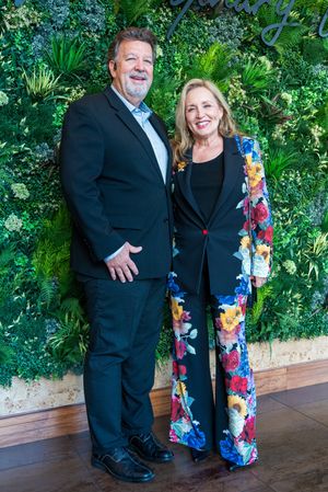 Two guests pose in formal attire against a lush green wall backdrop at an event.