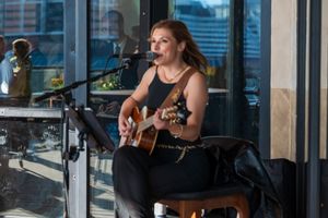 Acoustic performer plays guitar by large window in intimate venue setting.