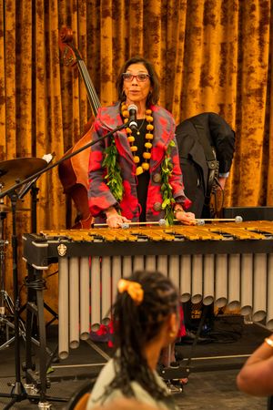 Musician wearing a lei performs behind a vibraphone marimba setup with golden curtains in the background.