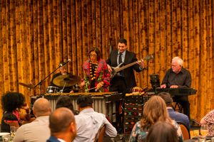 Band performing on stage with gold curtain backdrop at indoor event.