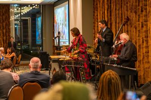 Musicians perform on stage with instruments including bass and drums against decorative curtains at an evening event.