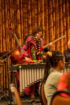 Musician in a vibrant red dress performs on percussion instruments at a concert with bamboo wall backdrop.