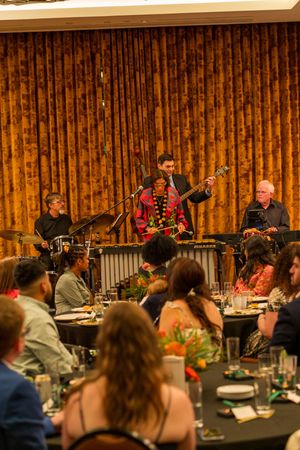 Musicians perform on stage with percussion instruments at an elegant dinner event with orange curtain backdrop.