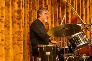 Drummer performing on a drum kit in front of gold curtains during a live performance.