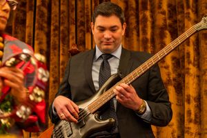 Bass guitarist in a dark suit performs on stage against golden curtains during an event.
