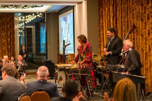 Jazz band performs live on stage at an elegant venue with performers playing drums and other instruments for seated audience.