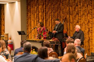 Musicians perform on stage with wood paneling backdrop while audience members watch in an intimate venue setting.