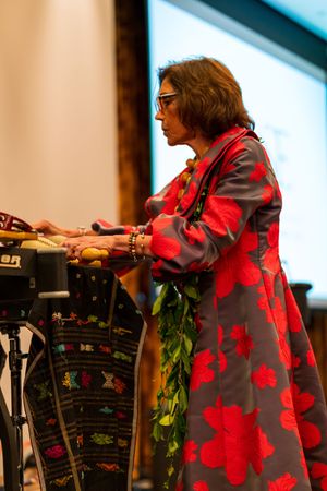 Musician in a red floral dress plays keyboard instruments on stage with tropical leaf decorations.