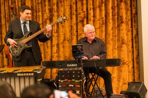 Band performers with various instruments including keyboard and guitar against golden curtain backdrop.