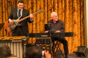 Musicians perform with bass guitar and keyboard against gold curtain backdrop on stage.