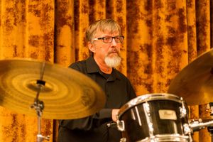 Drummer performing with cymbals and drum kit against orange curtain backdrop.
