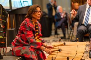 Musician in red plaid shirt performing with percussion instruments on wooden stage.