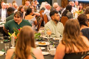 People seated at dining tables during a crowded banquet event with table settings and decorative flowers.