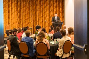 Speaker addressing audience members seated at round tables in a banquet hall.