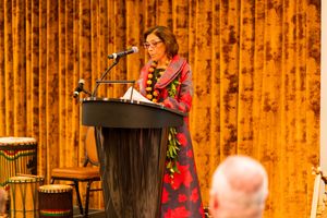 Speaker delivers presentation from wooden podium against gold curtain backdrop at formal event.